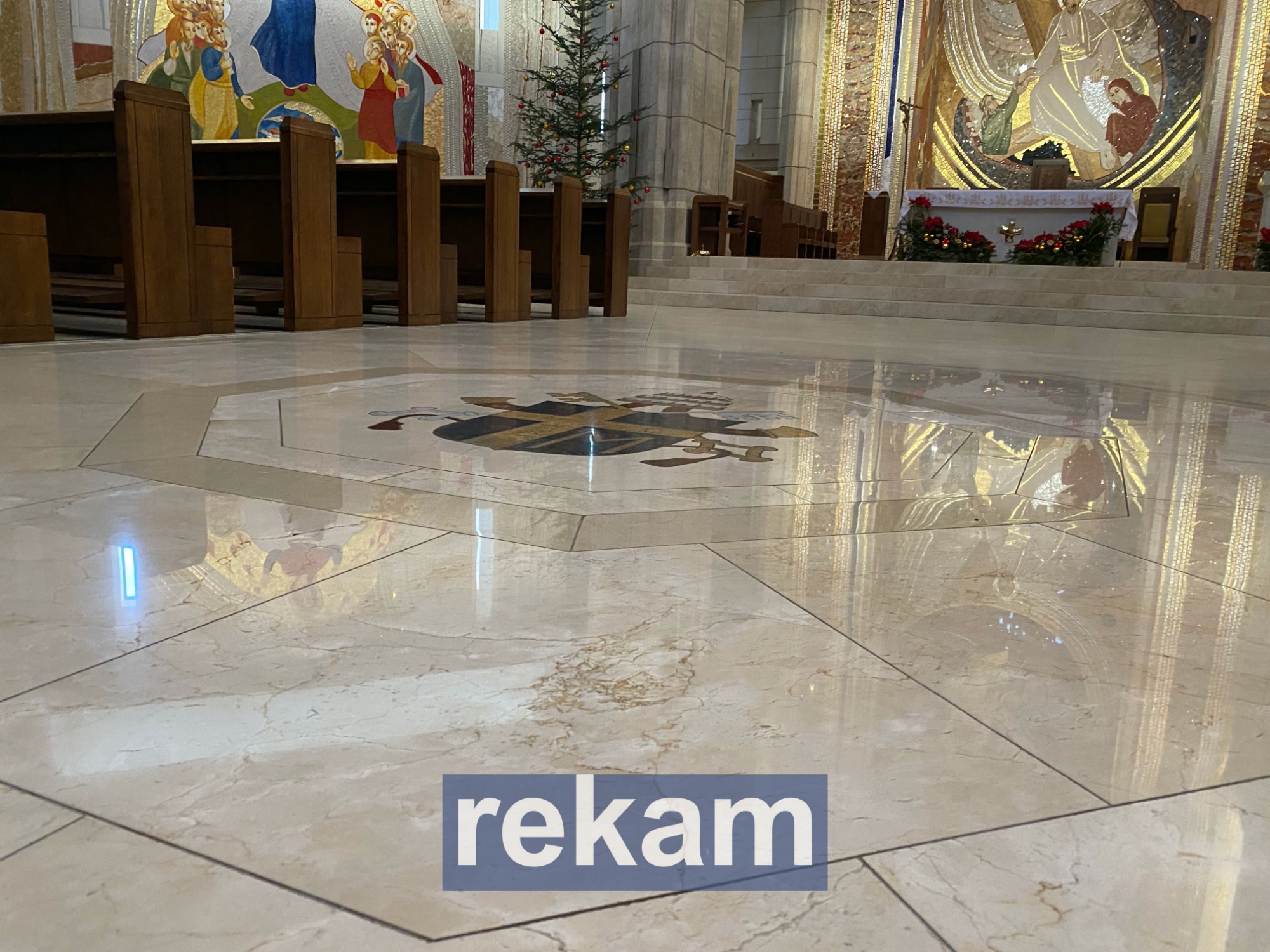 Jasna, marmurowa posadzka w kościele z herbem biskupim pośrodku, odbijająca oświetlenie i elementy wystroju wnętrza.
