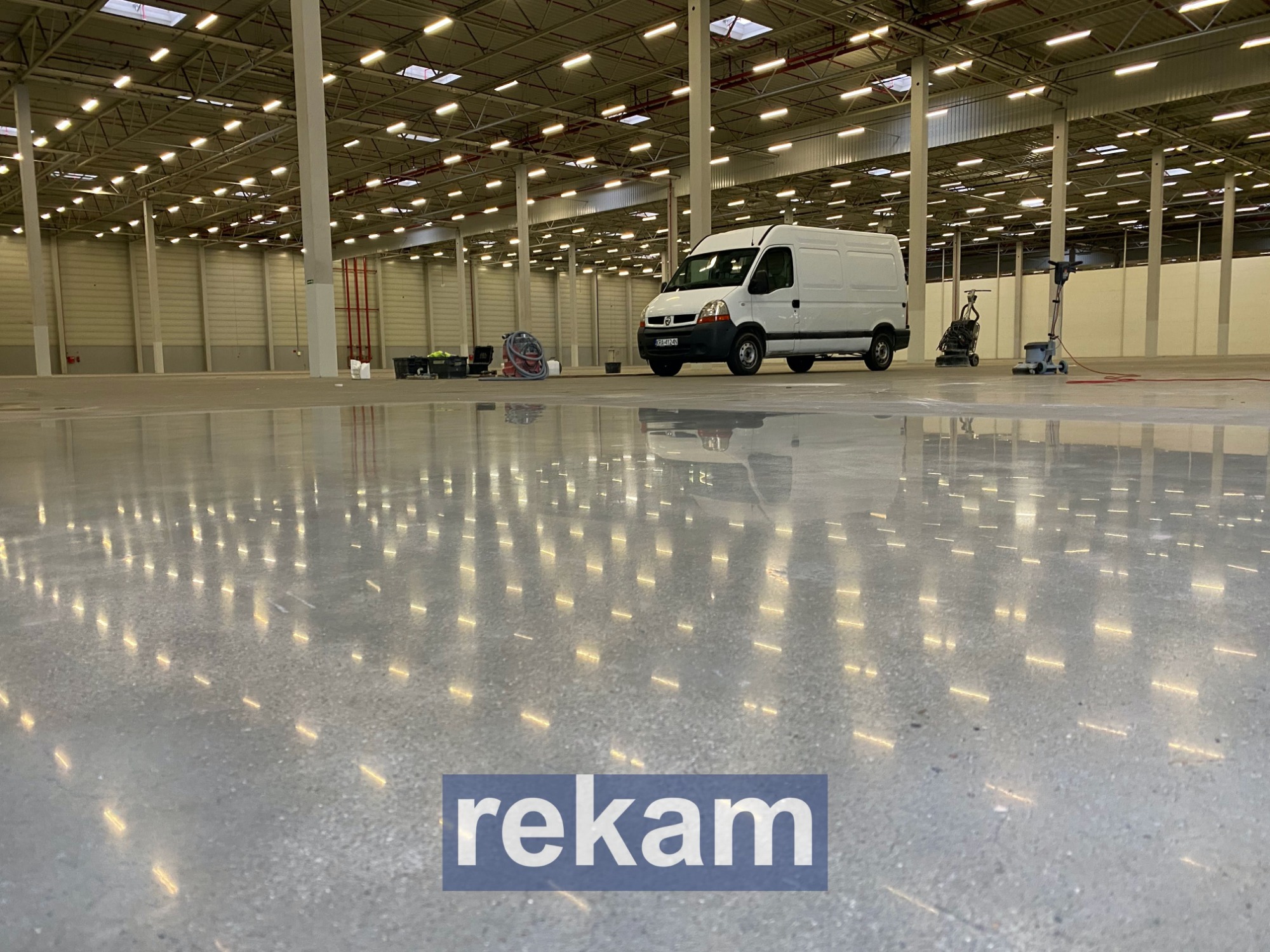 Błyszcząca, szara posadzka w hali magazynowej z odbijającymi się światłami, biały samochód dostawczy w tle oraz maszyny do czyszczenia posadzek.