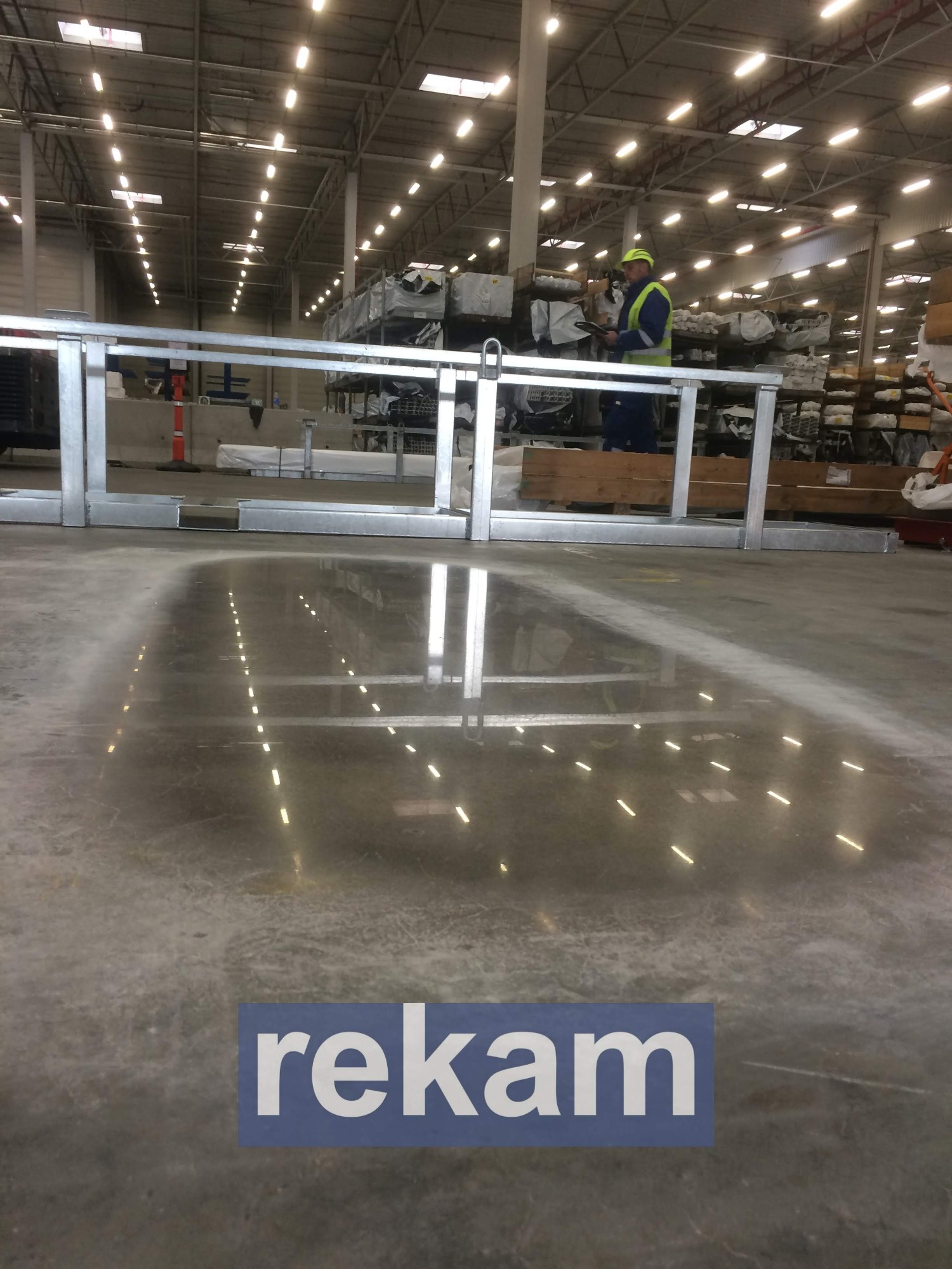 Błyszcząca, szara podłoga w hali magazynowej z odbiciami świateł, pracownik w odblaskowej kamizelce stoi w tle, widoczna metalowa konstrukcja i logo 'rekam' w dolnej części kadru.