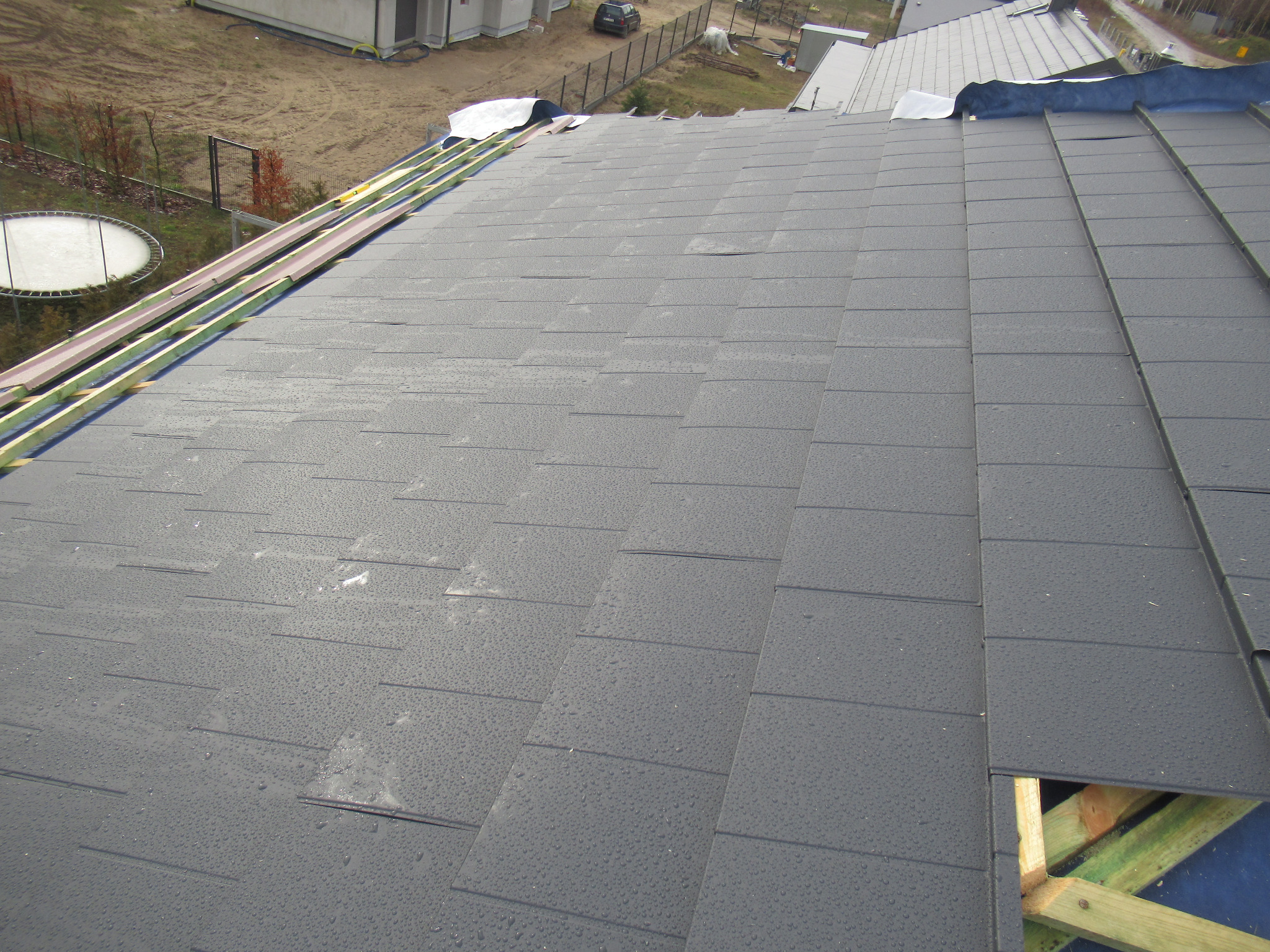 Montaż grafitowej dachówki na drewnianej konstrukcji, widoczne krople deszczu i elementy izolacji termicznej.