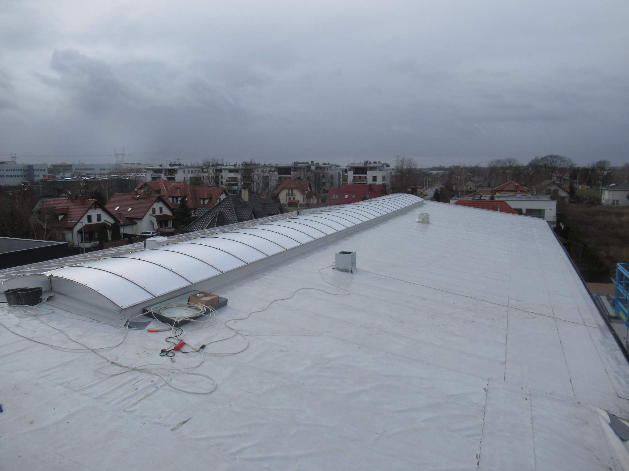 Biały dach płaski z świetlikami łukowymi, widoczne narzędzia i kable, w tle zabudowa miejska i pochmurne niebo.