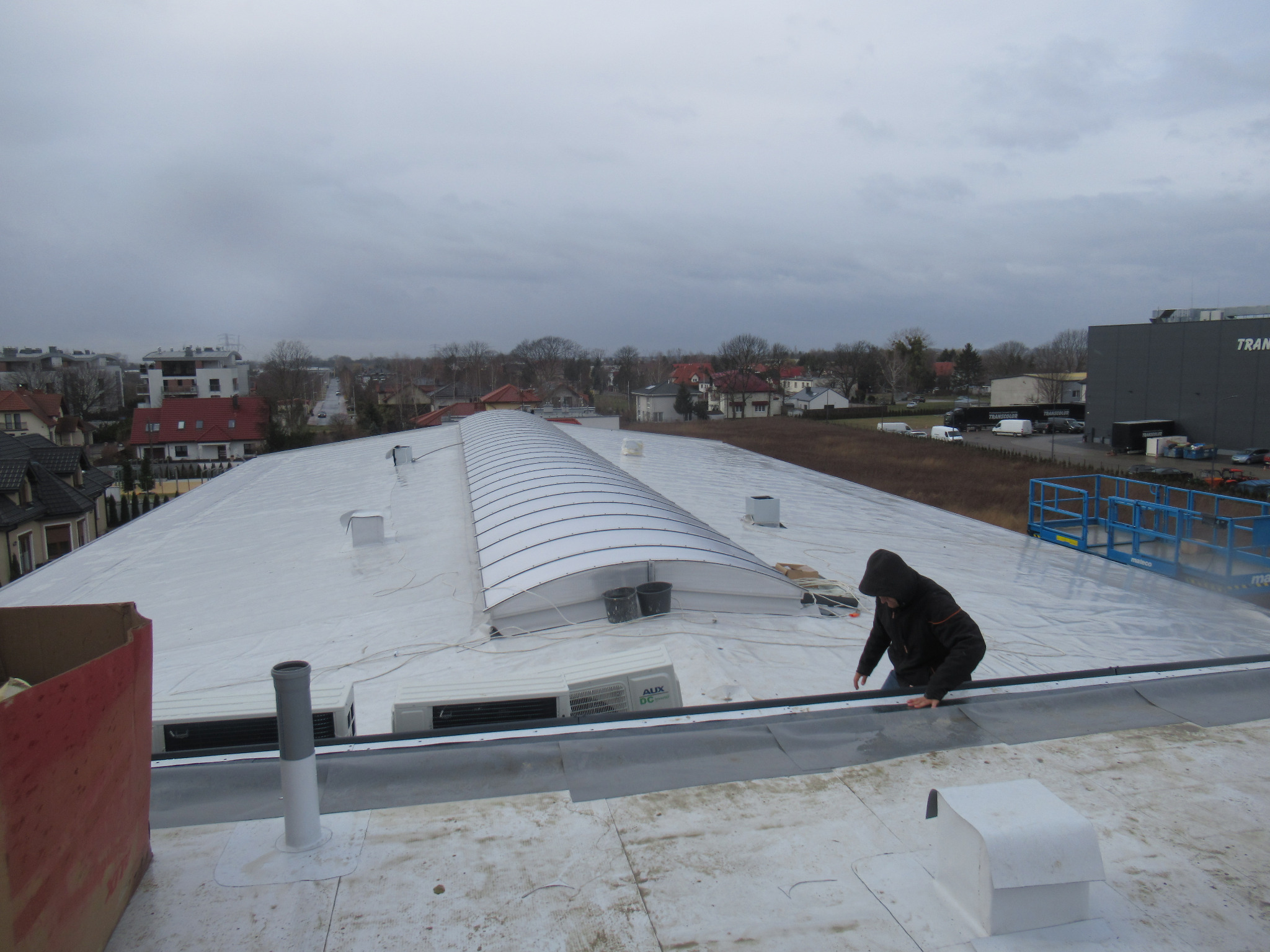 Pracownik w ciemnej kurtce z kapturem instaluje membranę na dachu hali przemysłowej z świetlikami, widoczny system klimatyzacji i budynki w tle.