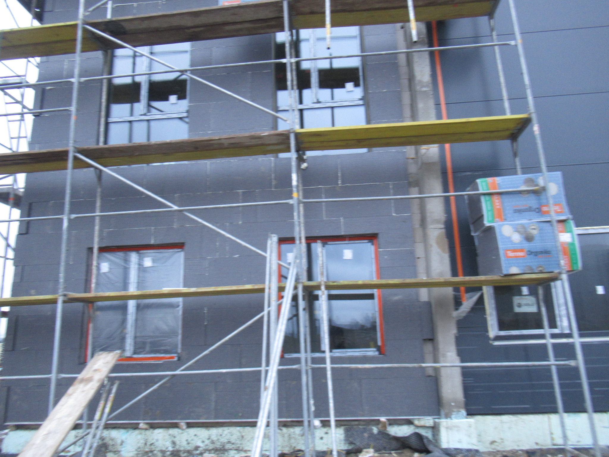 Budynek w trakcie ocieplania, widoczne rusztowania, okna z folią zabezpieczającą oraz paczki styropianu na elewacji.