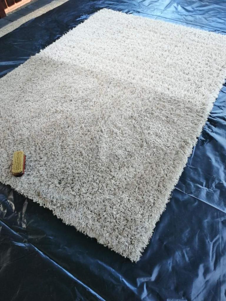 Beżowy dywan typu shaggy leżący na czarnej folii ochronnej, obok widoczna szczotka do czyszczenia.