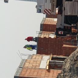 ArkadiaSp. z o.o. - Dwóch robotników w kaskach i odblaskowych kamizelkach pracuje na szczycie wznoszonego budynku z czerwonej cegły, widoczny dźwig budowlany w tle.