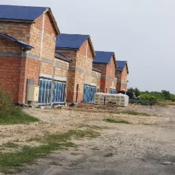 Szereg budynków w stanie surowym otwartym z niebieskim pokryciem dachowym, widoczne okna zabezpieczone folią, materiały budowlane składowane na zewnątrz, mężczyzna przechodzący obok.