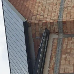 Fragment budynku w trakcie budowy: widoczna ściana z cegieł, fragment dachu z blachy trapezowej i zamontowana rynna.