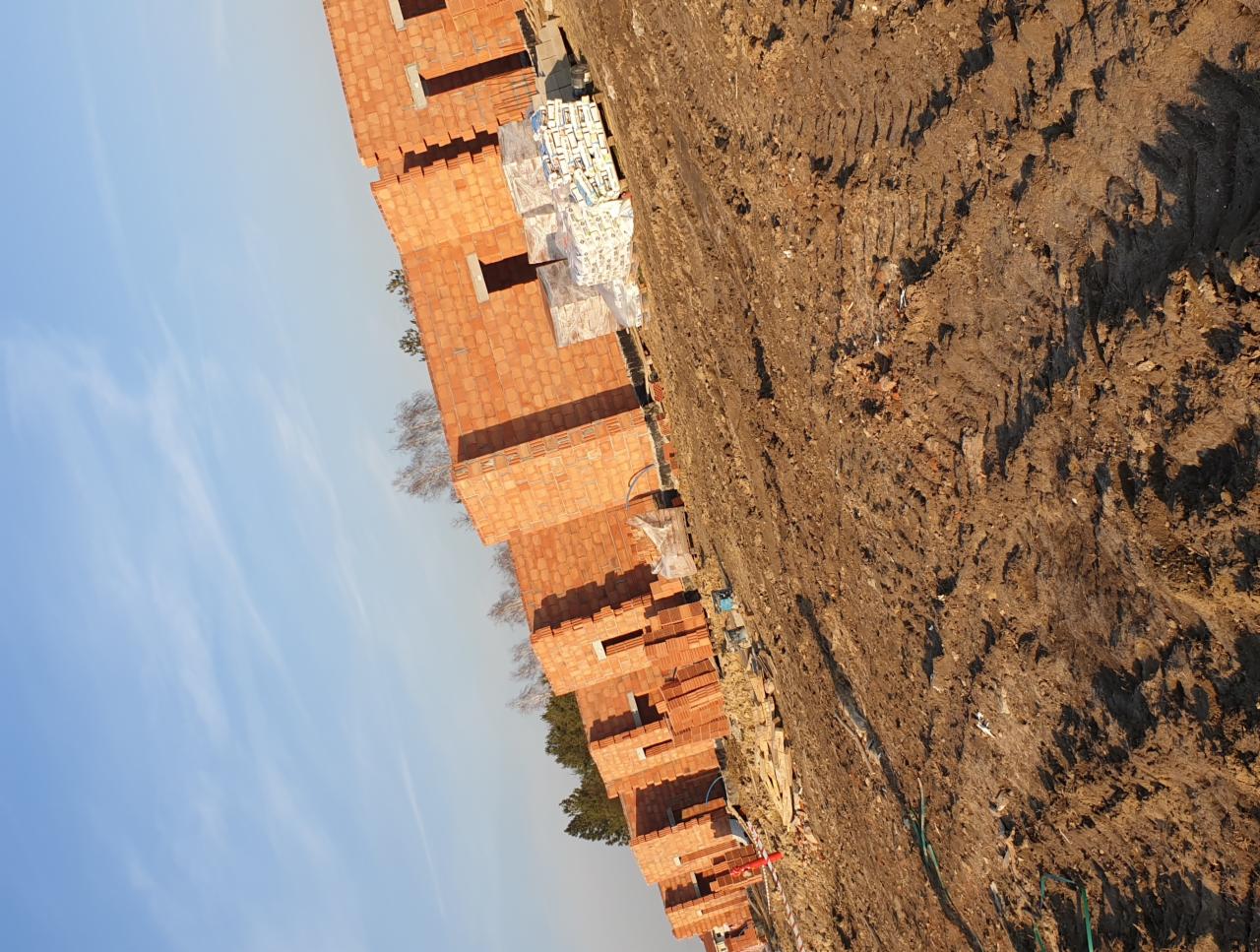 Wczesny etap budowy domu z czerwonej cegły ceramicznej, widoczne pakiety materiałów budowlanych, przygotowany teren z widocznymi śladami pojazdów budowlanych.
