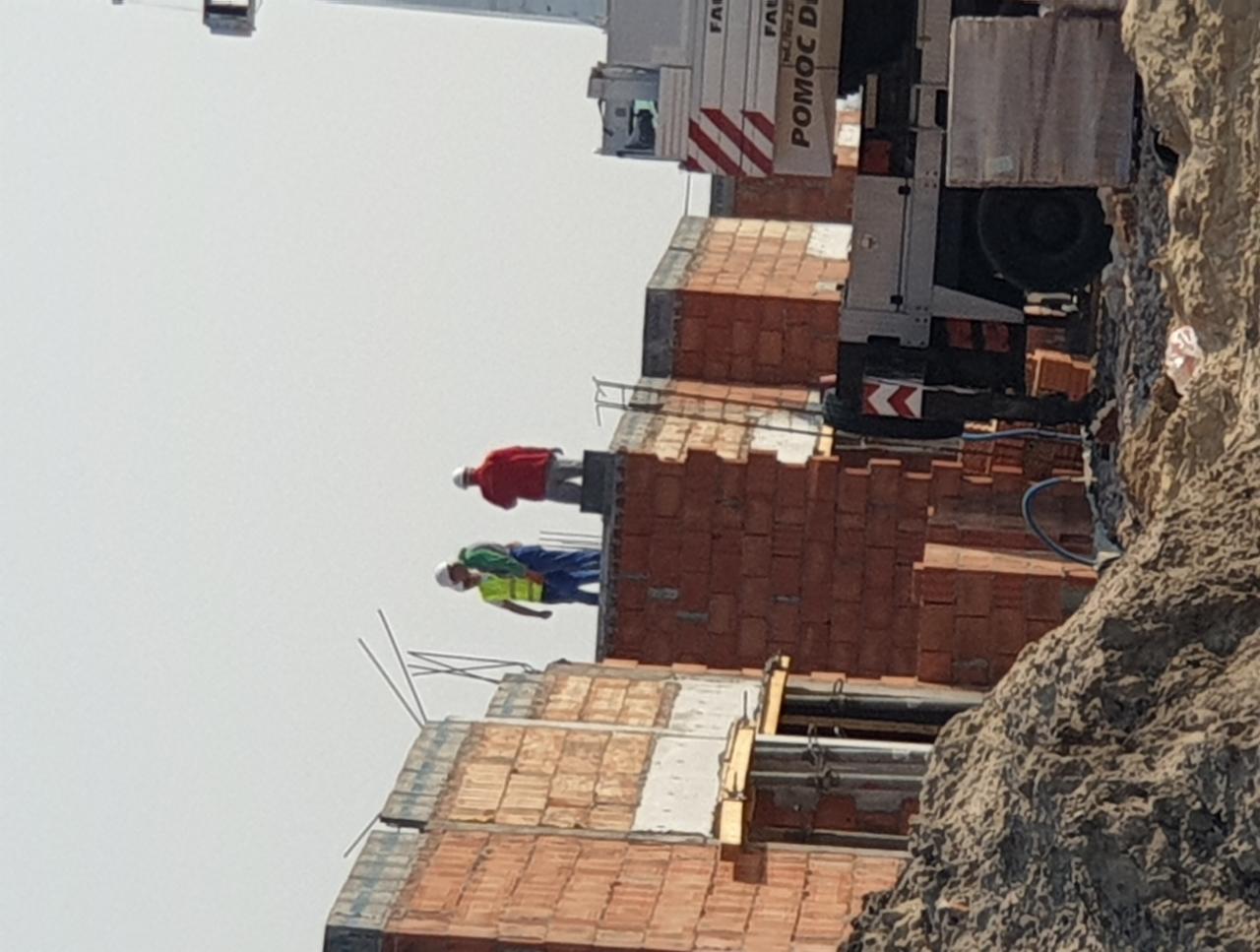 Dwóch robotników w kaskach i odblaskowych kamizelkach pracuje na szczycie wznoszonego budynku z czerwonej cegły, widoczny dźwig budowlany w tle.