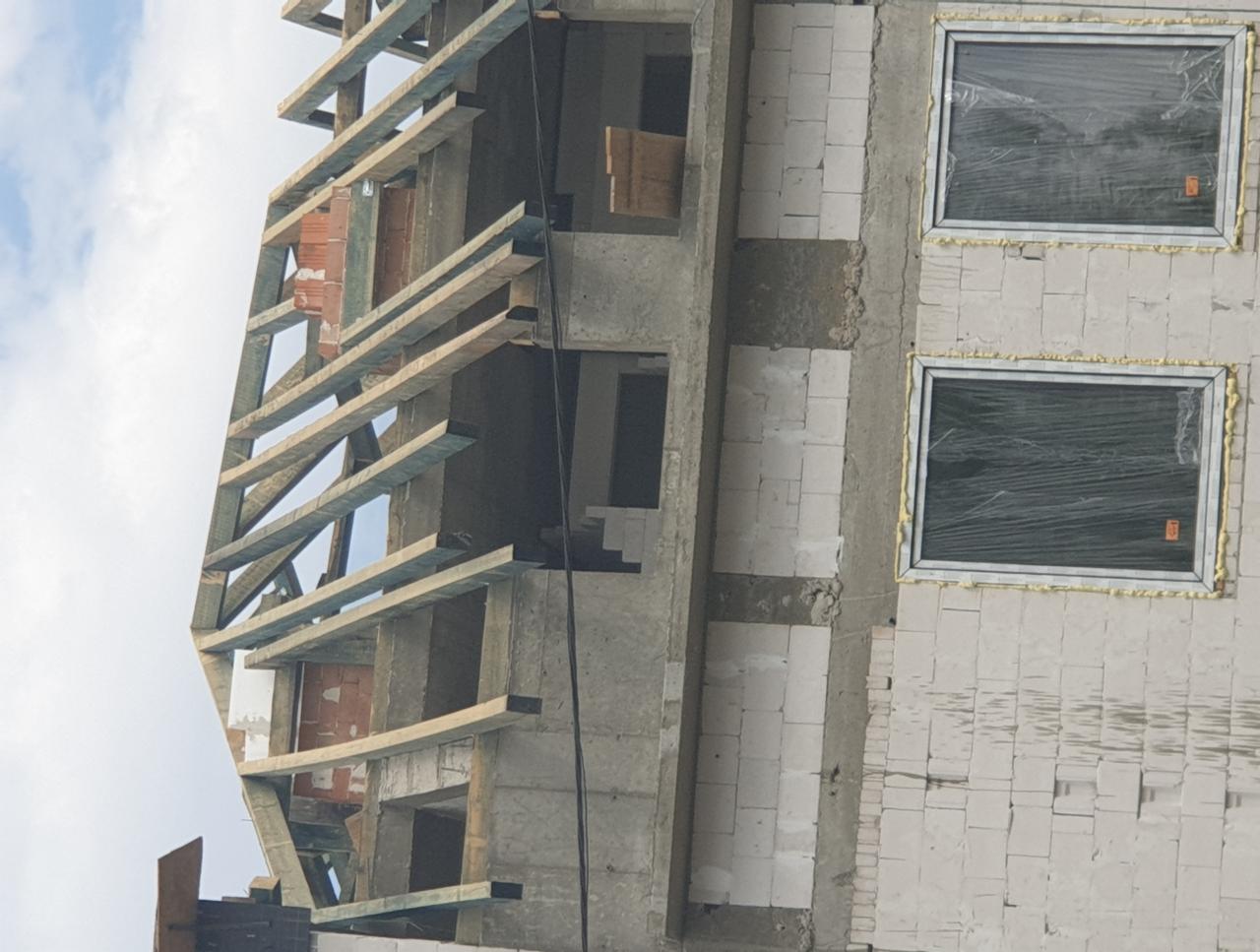 Surowy stan budynku wielokondygnacyjnego w trakcie budowy, widoczne białe bloczki, drewniane szalunki i okna zabezpieczone folią.