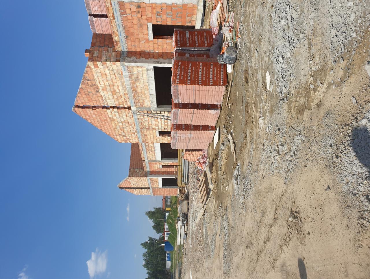 Wznoszony budynek z czerwonej cegły Termoton, widok z poziomu gruntu na tle błękitnego nieba, palety cegieł obok fundamentów.