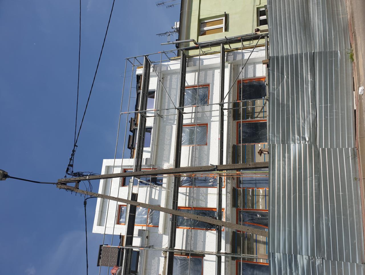 Budynek wielorodzinny w trakcie ocieplania, widoczne rusztowania, zabezpieczone okna folią i blacha trapezowa osłaniająca fragment elewacji.