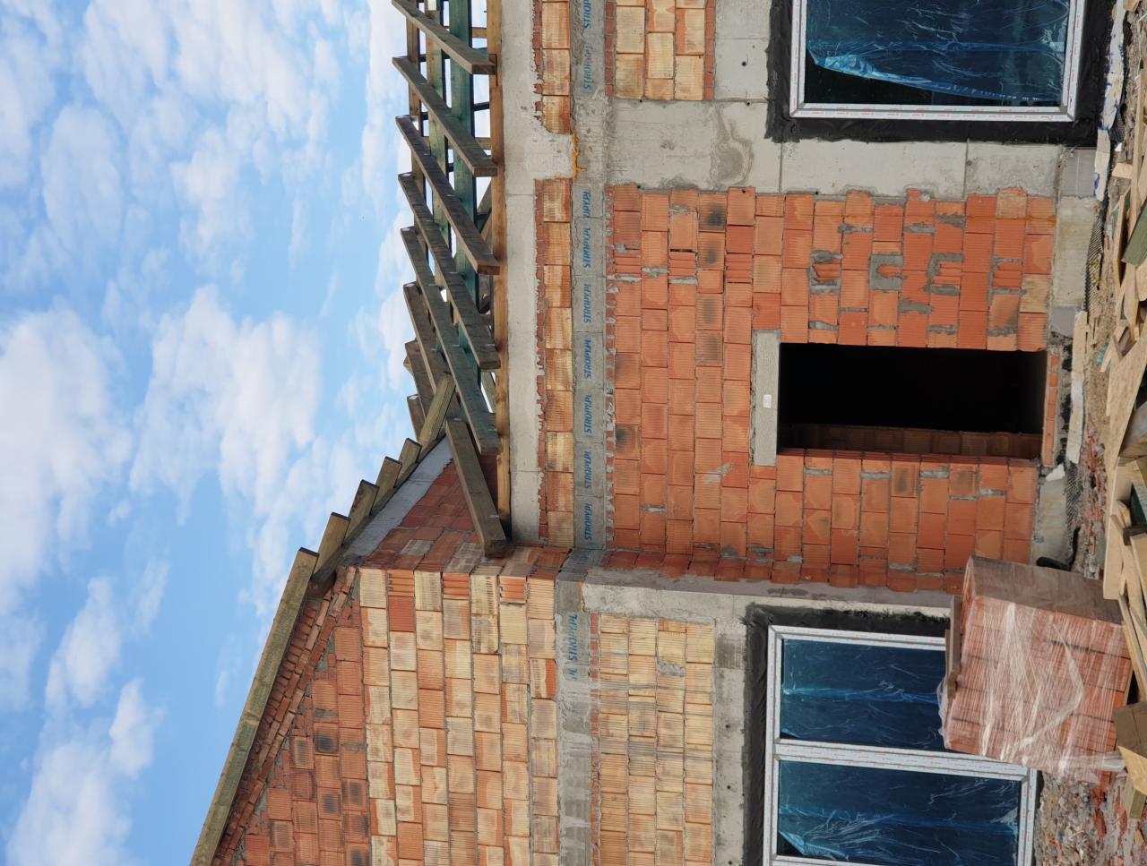 Wznoszone ściany domu z czerwonej cegły z widocznymi oknami zabezpieczonymi folią i konstrukcją dachu w trakcie budowy na tle błękitnego nieba.