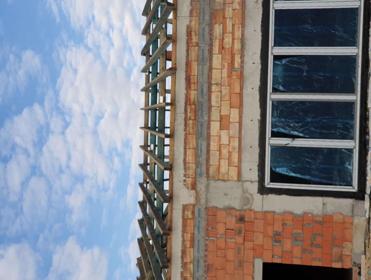 Wznoszona ściana z czerwonej cegły z oknem zabezpieczonym folią, widoczne drewniane elementy konstrukcji dachu na tle błękitnego nieba z chmurami.