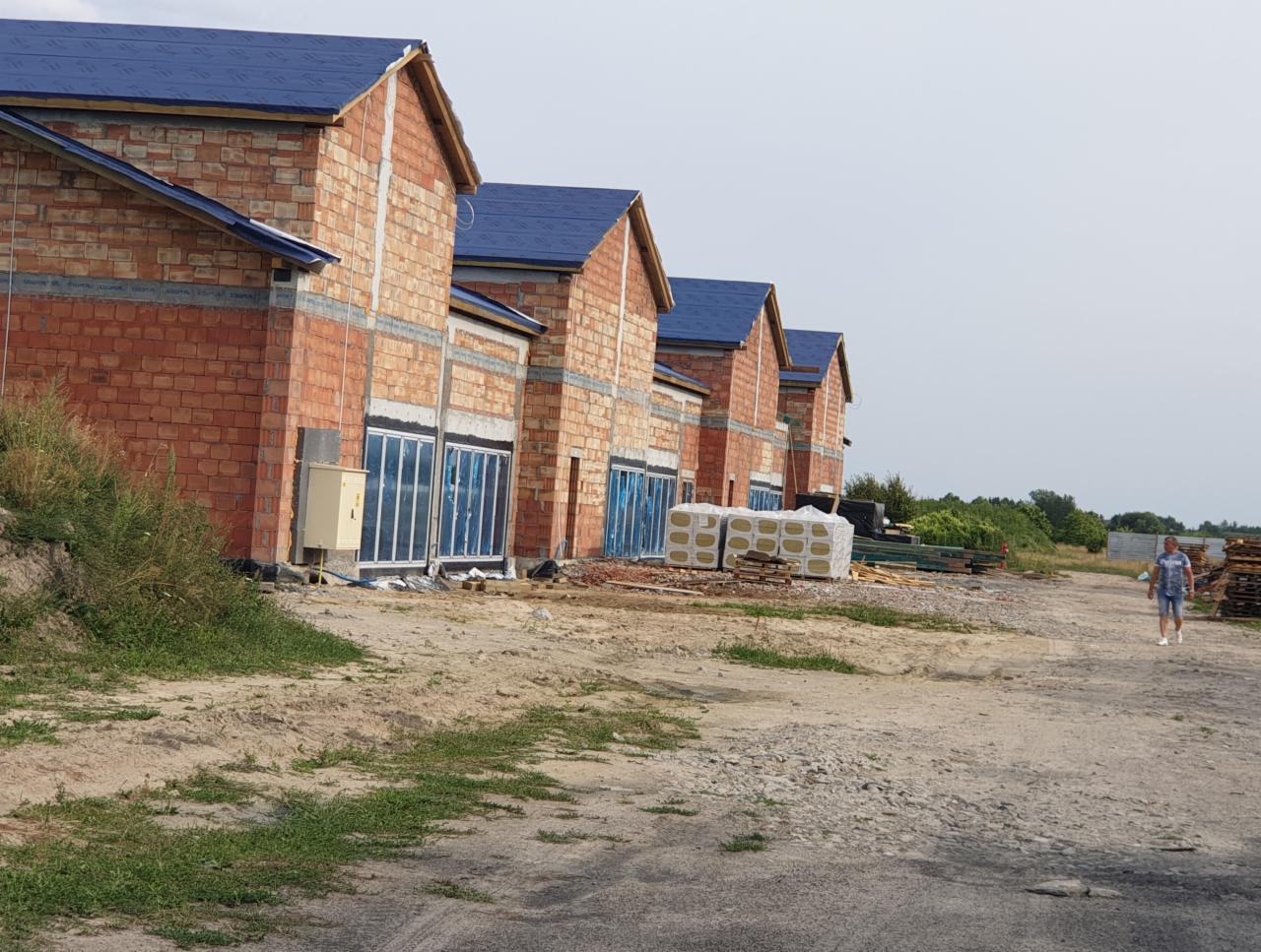 Szereg budynków w stanie surowym otwartym z niebieskim pokryciem dachowym, widoczne okna zabezpieczone folią, materiały budowlane składowane na zewnątrz, mężczyzna przechodzący obok.