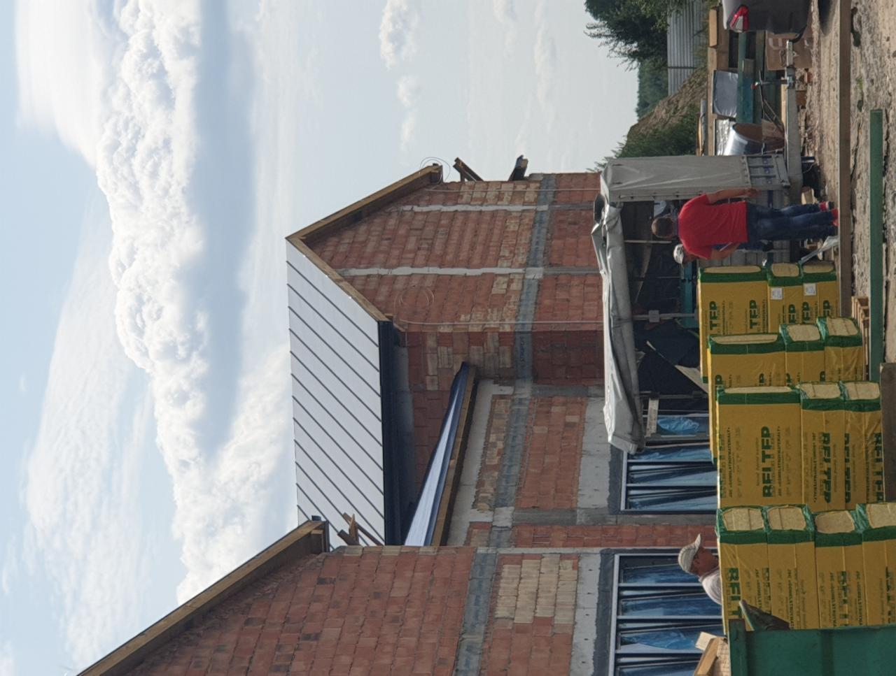 Budowa domu murowanego w trakcie realizacji, widoczne ściany z cegły, częściowo zamontowane okna zabezpieczone folią, blachy na dachu oraz palety z materiałami izolacyjnymi.