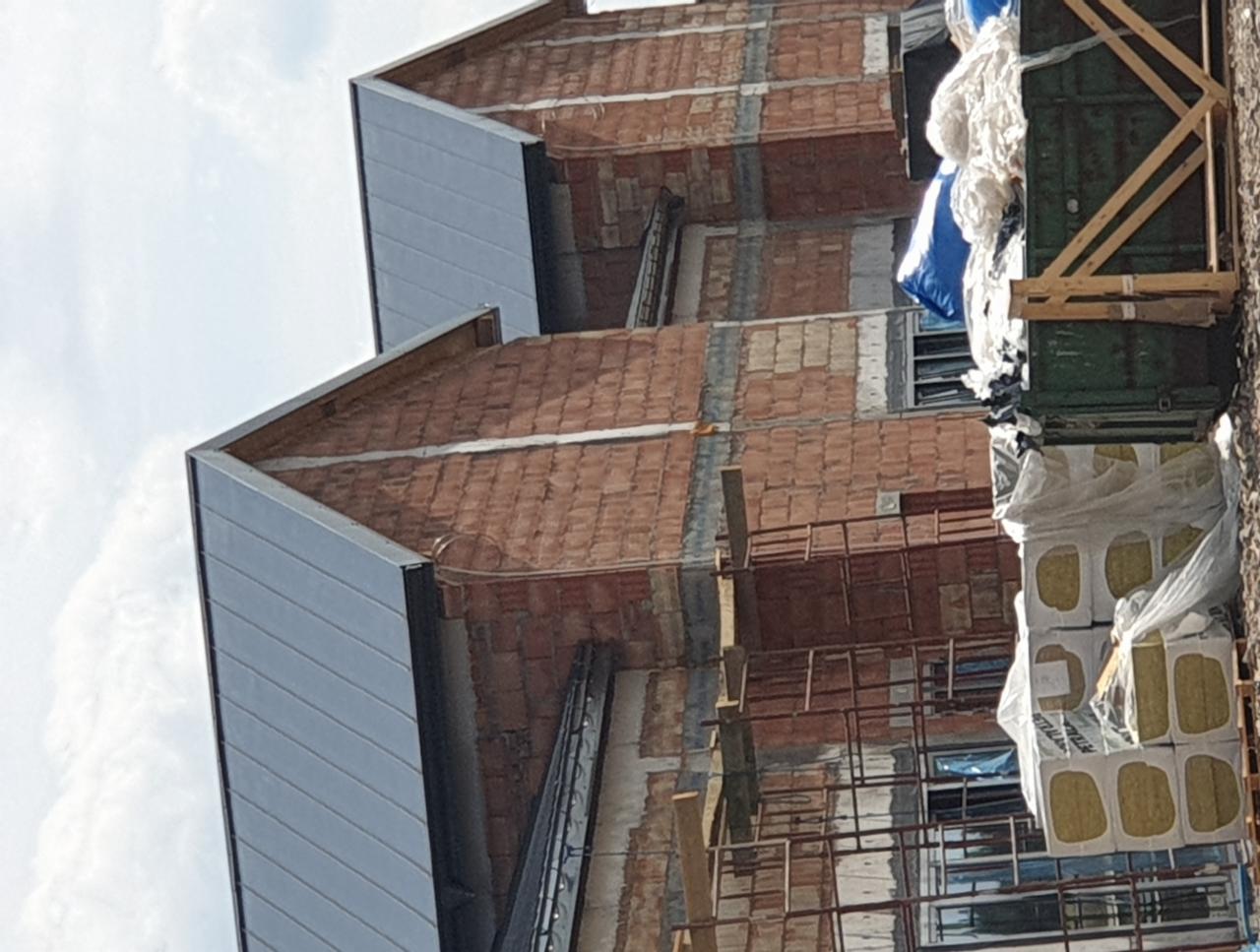 Surowy stan budowy domu z cegły z widocznym ociepleniem i fragmentem dachu z blachy na tle pochmurnego nieba.