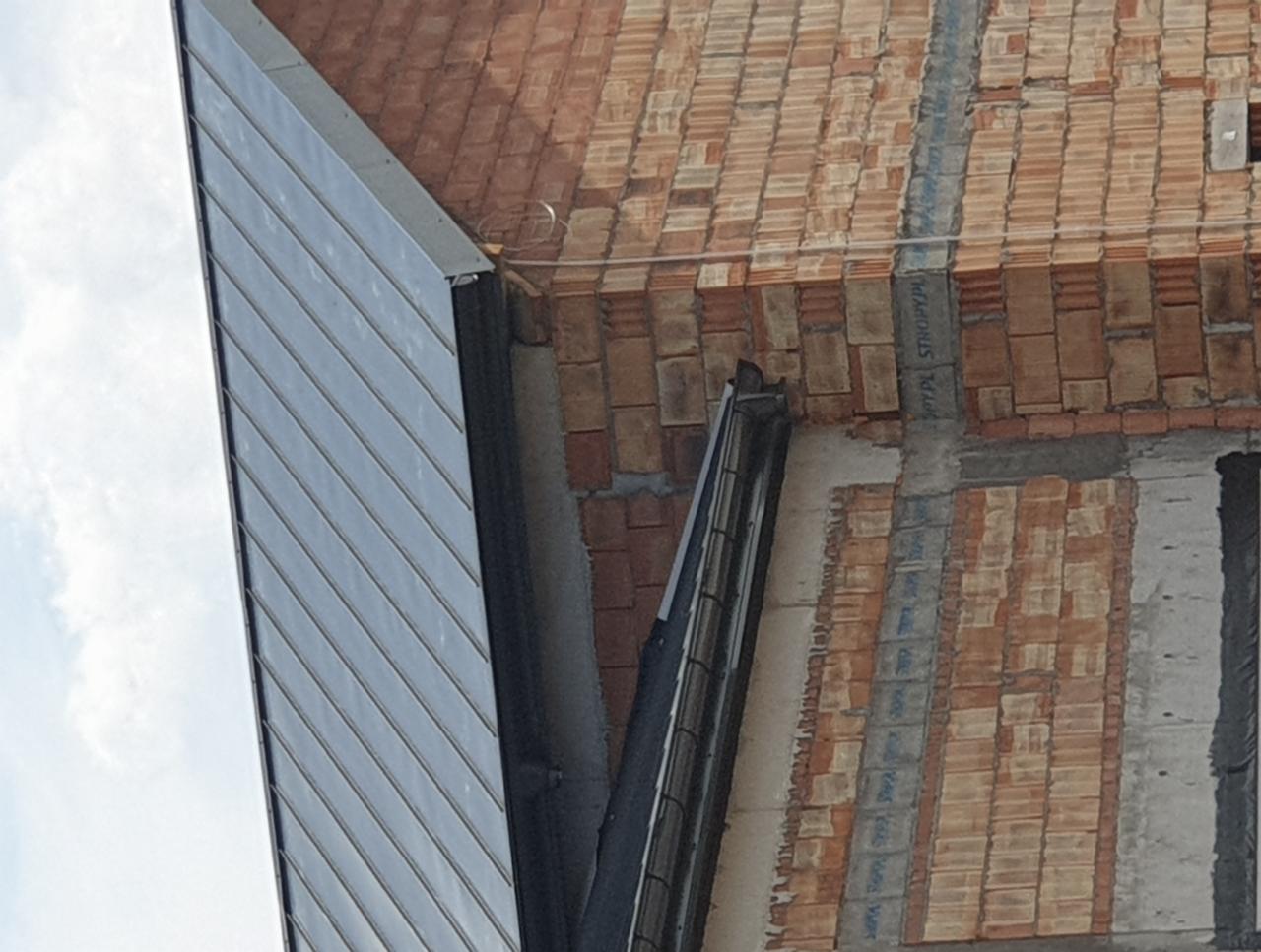 Fragment budynku w trakcie budowy: widoczna ściana z cegieł, fragment dachu z blachy trapezowej i zamontowana rynna.