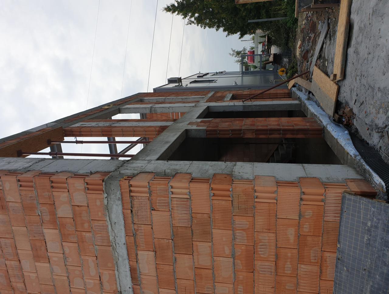 Wznoszenie ścian z czerwonej cegły na konstrukcji szkieletowej, widoczne zbrojenia betonowe i elementy drewniane więźby dachowej w surowym stanie.