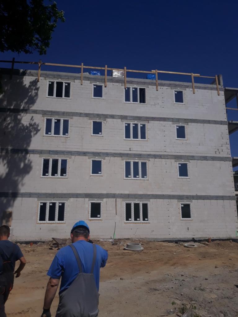 Wznoszony, trzypiętrowy budynek z betonu komórkowego z zamontowanymi oknami, widoczne szalunki stropowe na dachu, teren budowy z dwoma robotnikami w tle.