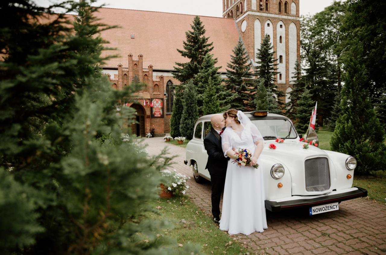 Pocałunek nowożeńców na tle ceglanego kościoła i białej taksówki z kwiatami, tablica rejestracyjna z napisem 'Nowożeńcy'.