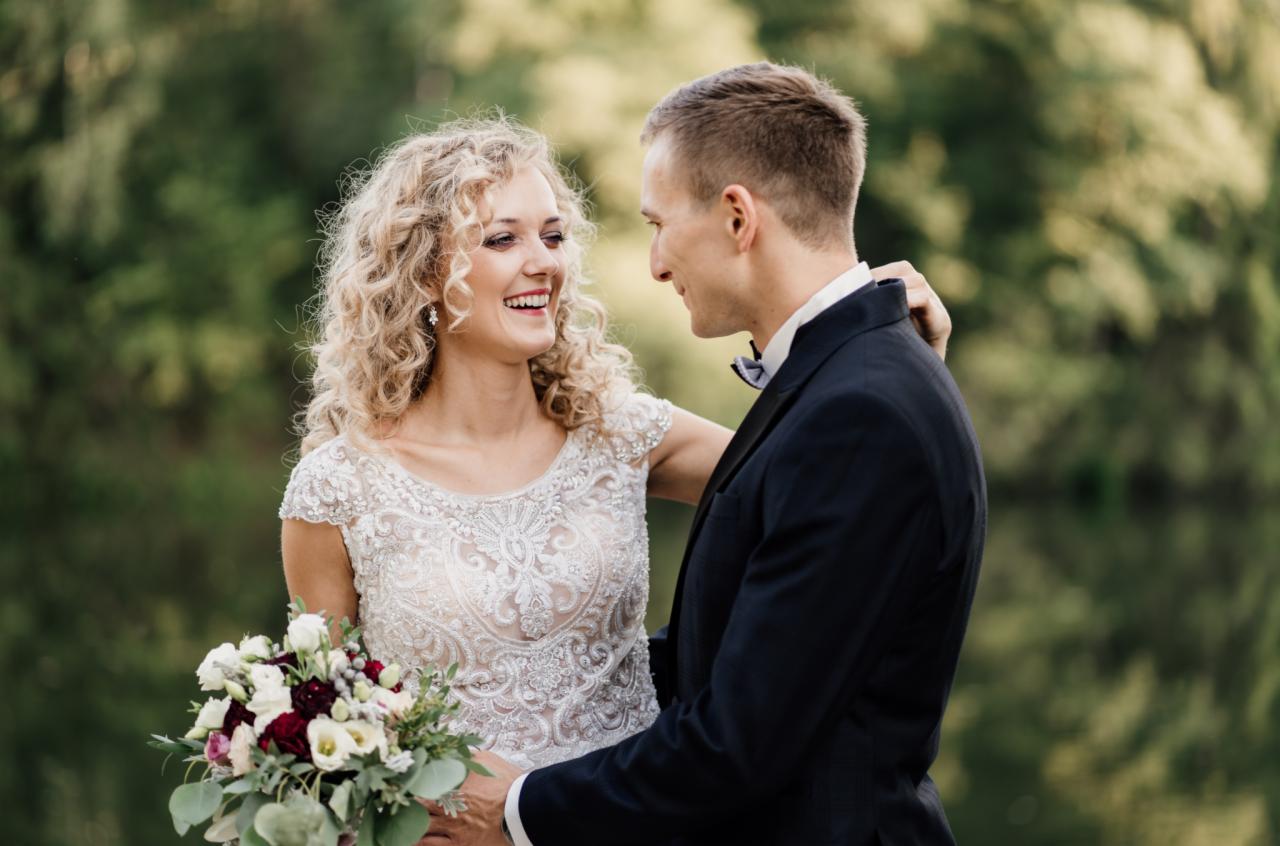 Uśmiechnięta panna młoda w koronkowej sukni i z bukietem, obejmowana przez pana młodego w ciemnym garniturze z muchą, na tle zieleni.