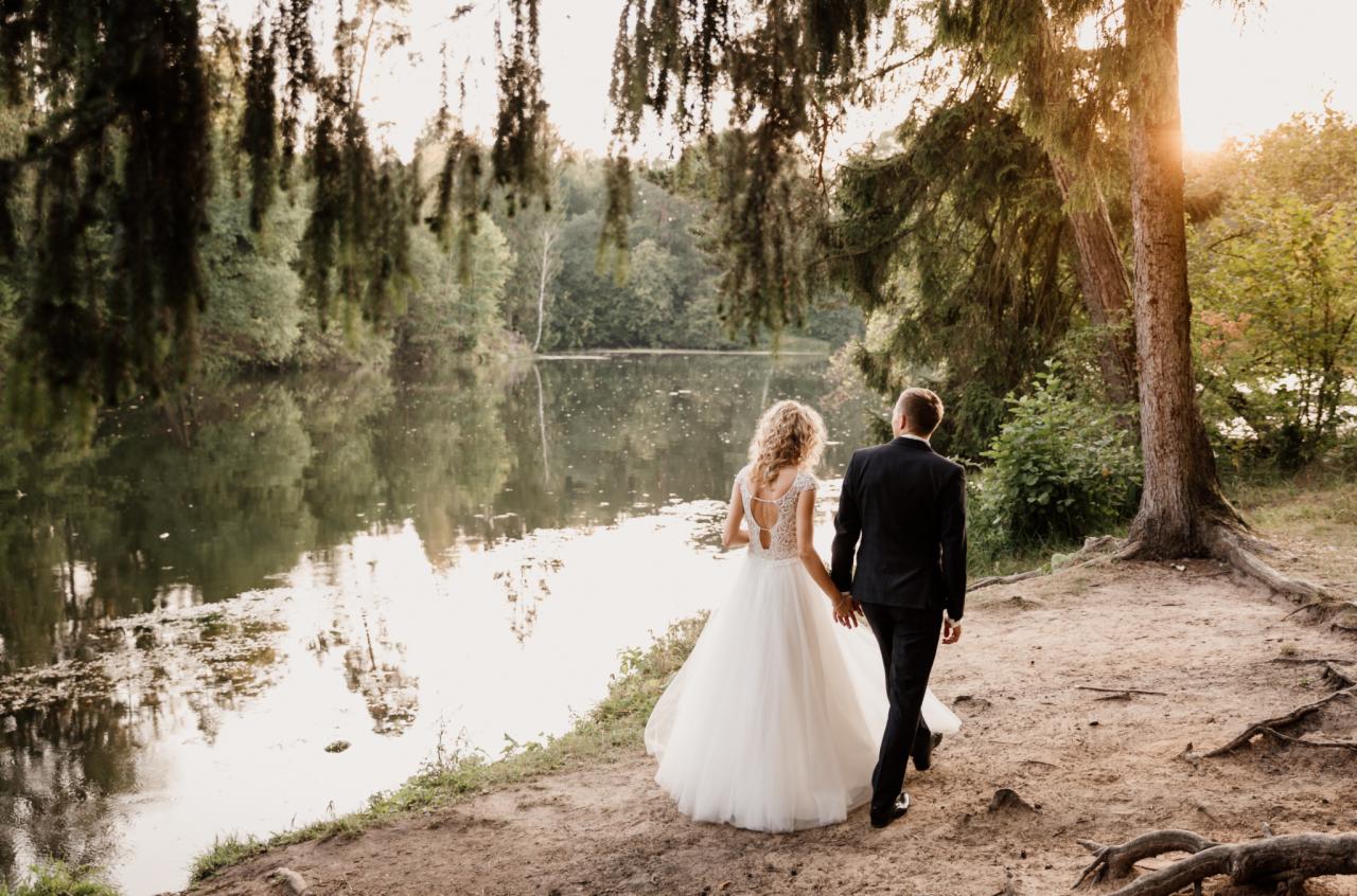 Para młoda, on w czarnym garniturze, ona w białej sukni z koronkowymi plecami, spaceruje brzegiem jeziora w lesie, trzymając się za ręce w promieniach zachodzącego słońca.