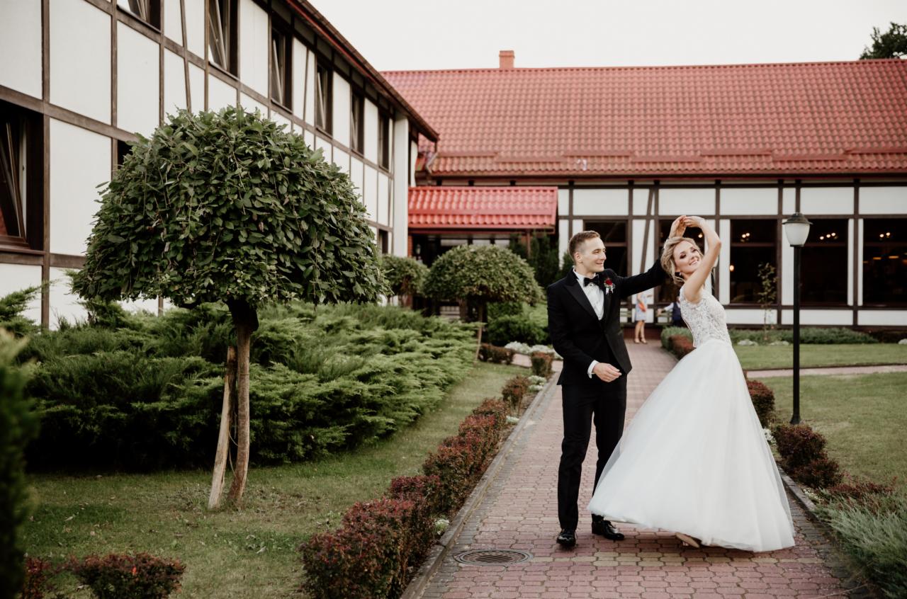 Uśmiechnięta panna młoda w koronkowej sukni i pan młody w smokingu tańczą na brukowanej ścieżce przed budynkiem z czerwonym dachem i białymi ścianami w kratkę.