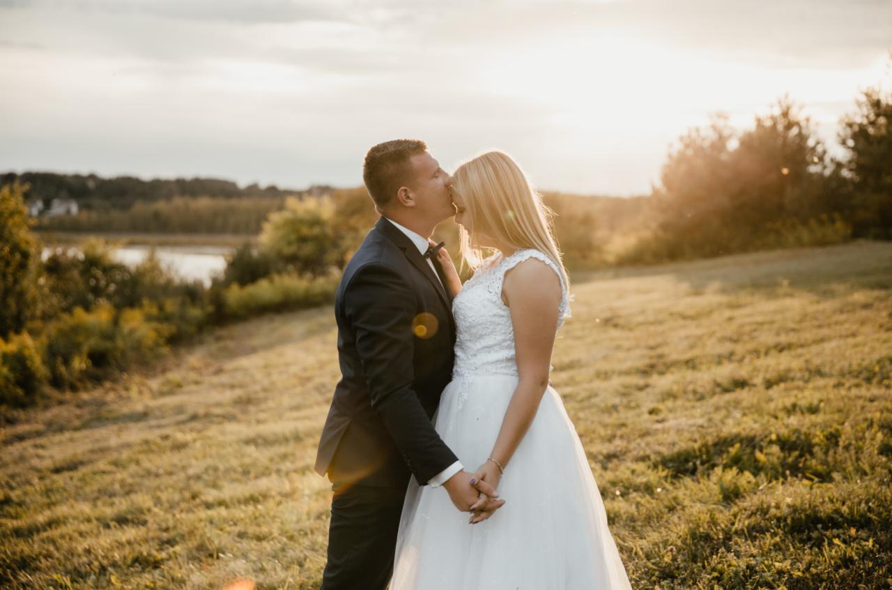 Para młoda w objęciach na łące o zachodzie słońca, on w ciemnym garniturze całuje ją w czoło, ona w białej sukni z koronkową górą, trzymają się za ręce.