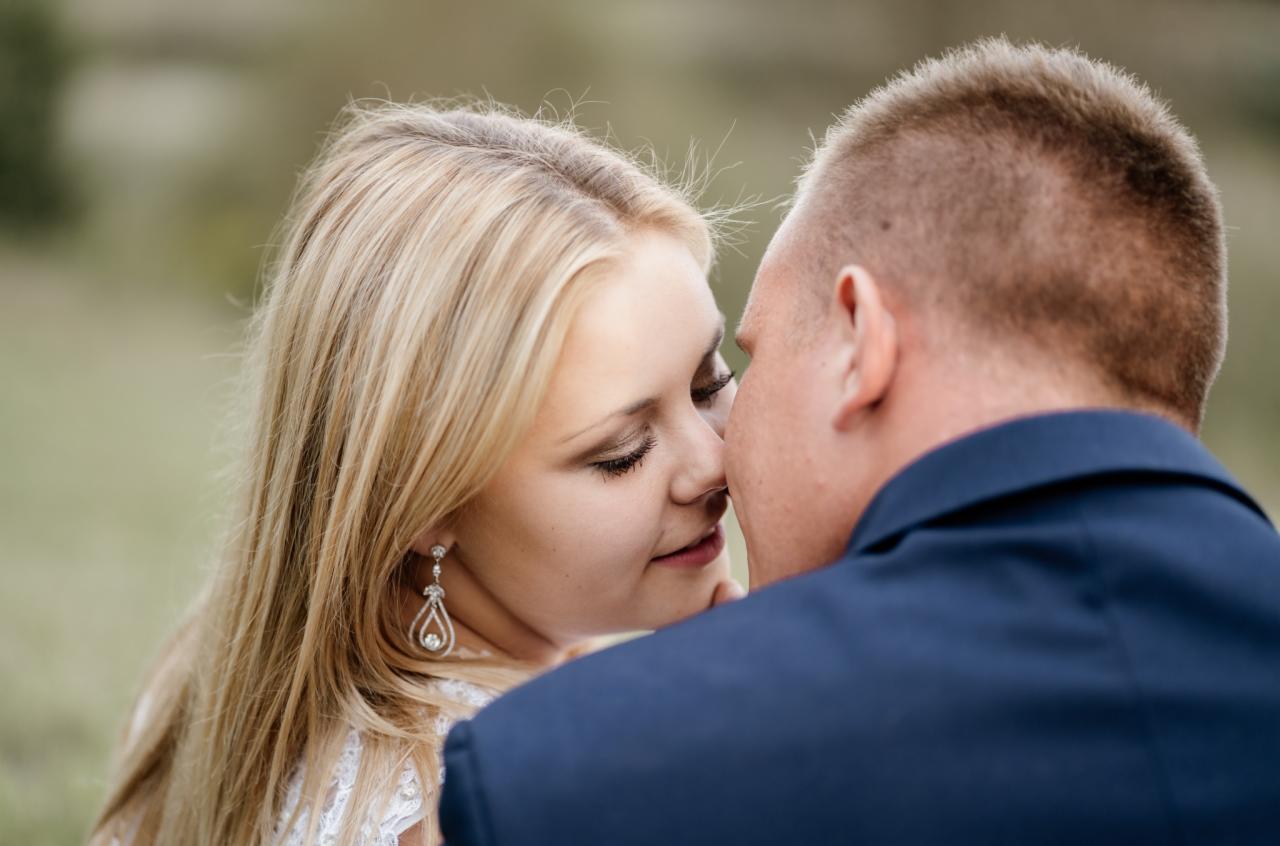 Panna młoda z blond włosami i wiszącymi kolczykami, delikatnie dotyka nosem policzka pana młodego w granatowym garniturze, na rozmytym, zielonym tle.