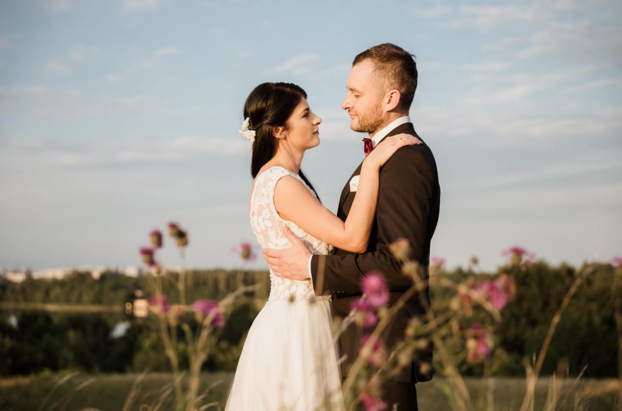Para młoda, brunetka w koronkowej sukni i brunet w czarnym garniturze z bordową muchą, obejmują się na tle łąki z fioletowymi kwiatami i błękitnego nieba.