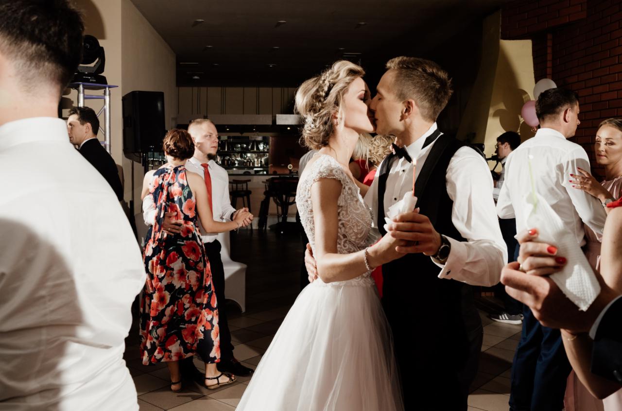 Pocałunek pary młodej podczas pierwszego tańca na tle gości weselnych i baru.