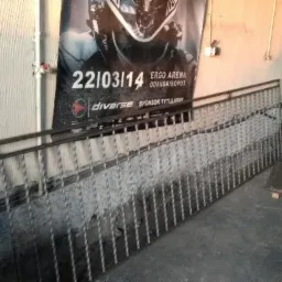 Fragment metalowej balustrady z dekoracyjnymi, skręconymi elementami pionowymi, oparta o ścianę z plakatem reklamowym w tle.