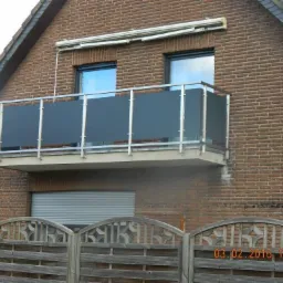 Balkon z szarą, pełną balustradą i srebrnymi poręczami, zamontowany na ceglanym budynku z widocznym kominem i drewnianym ogrodzeniem w tle.