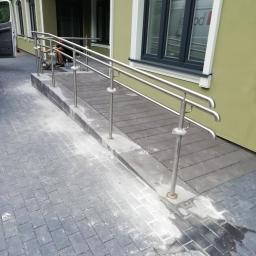 Bauservice - Zewnętrzna rampa z trzypoziomowymi poręczami ze stali nierdzewnej przed budynkiem, widoczne ślady po montażu na kostce brukowej, pracownik w tle.