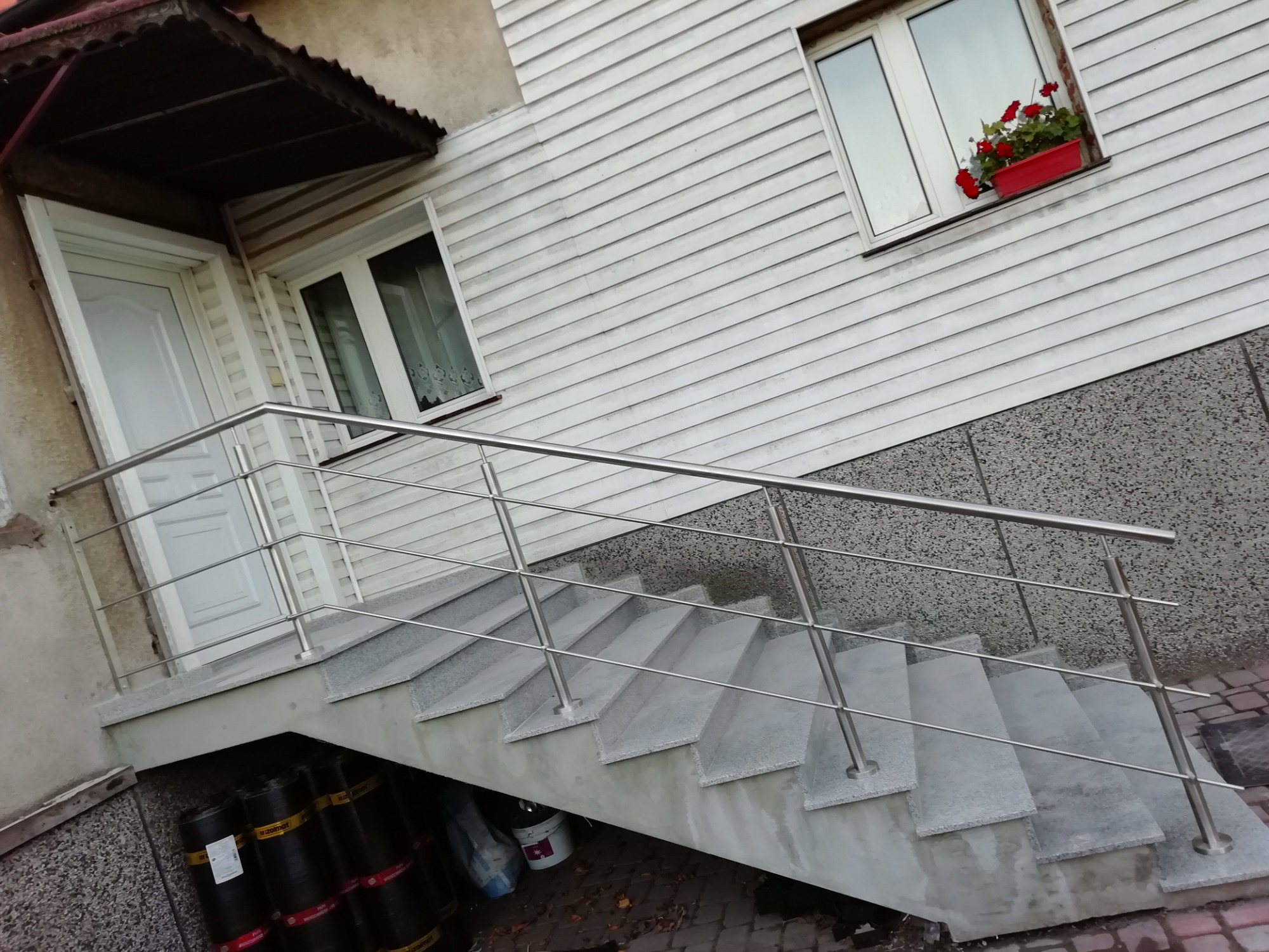 Zewnętrzne schody z szarego betonu z prostą, metalową poręczą, prowadzące do białych drzwi wejściowych. Pod schodami widoczne materiały budowlane, a w oknie na piętrze czerwone kwiaty.