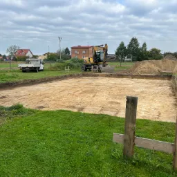 Przygotowany teren pod budowę domu: wykopany fundament, żółta koparka CAT, biała ciężarówka w tle, widoczne drewniane stemple pomiarowe.