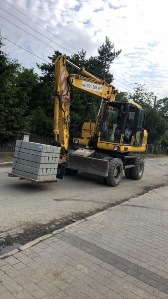 Żółta koparka kołowa przewozi paletę z szarymi bloczkami betonowymi na ulicy wyłożonej kostką. Na wysięgniku koparki widoczny numer telefonu. W tle drzewa i słupy energetyczne.