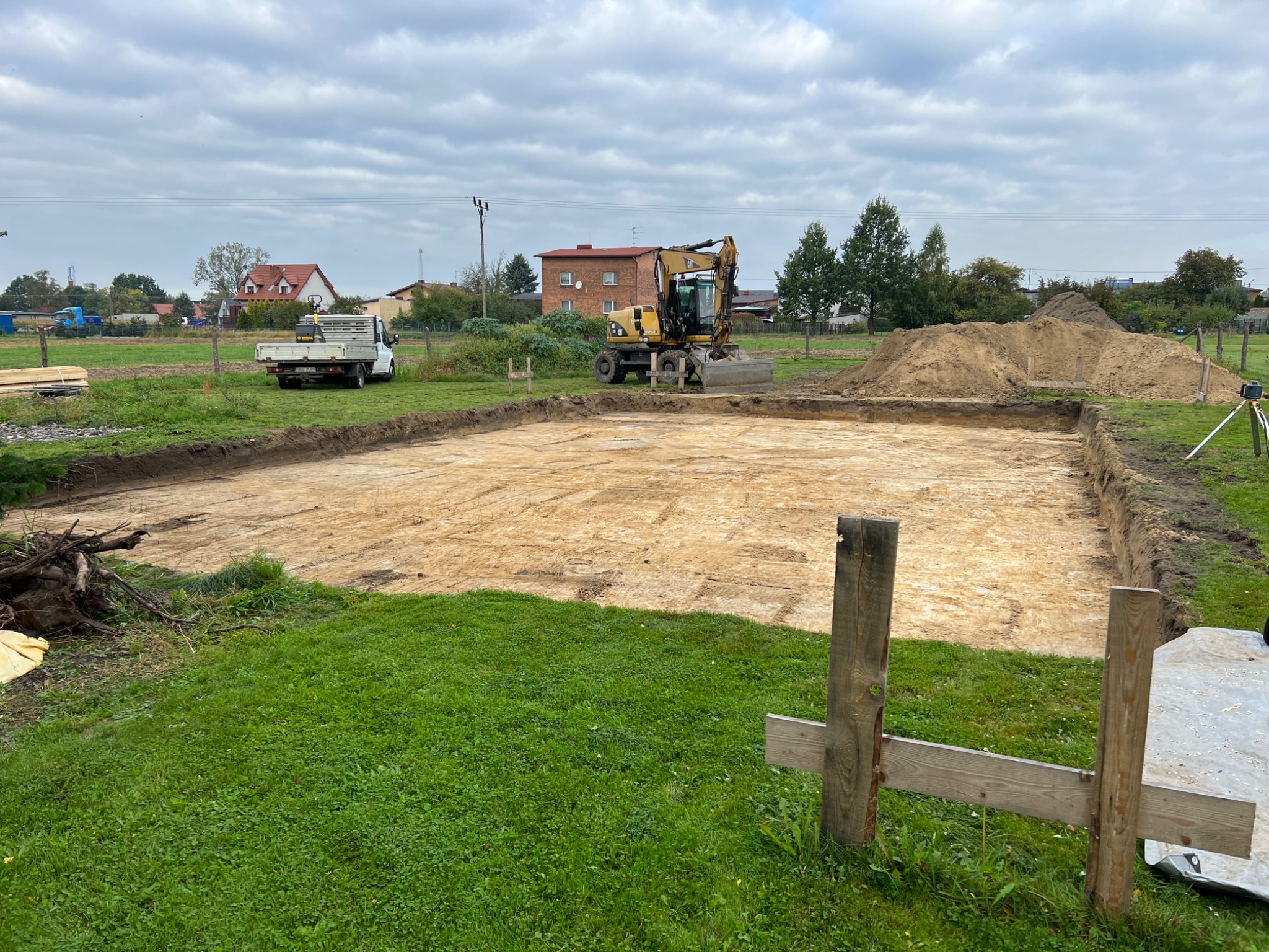 Przygotowany teren pod budowę domu: wykopany fundament, żółta koparka CAT, biała ciężarówka w tle, widoczne drewniane stemple pomiarowe.