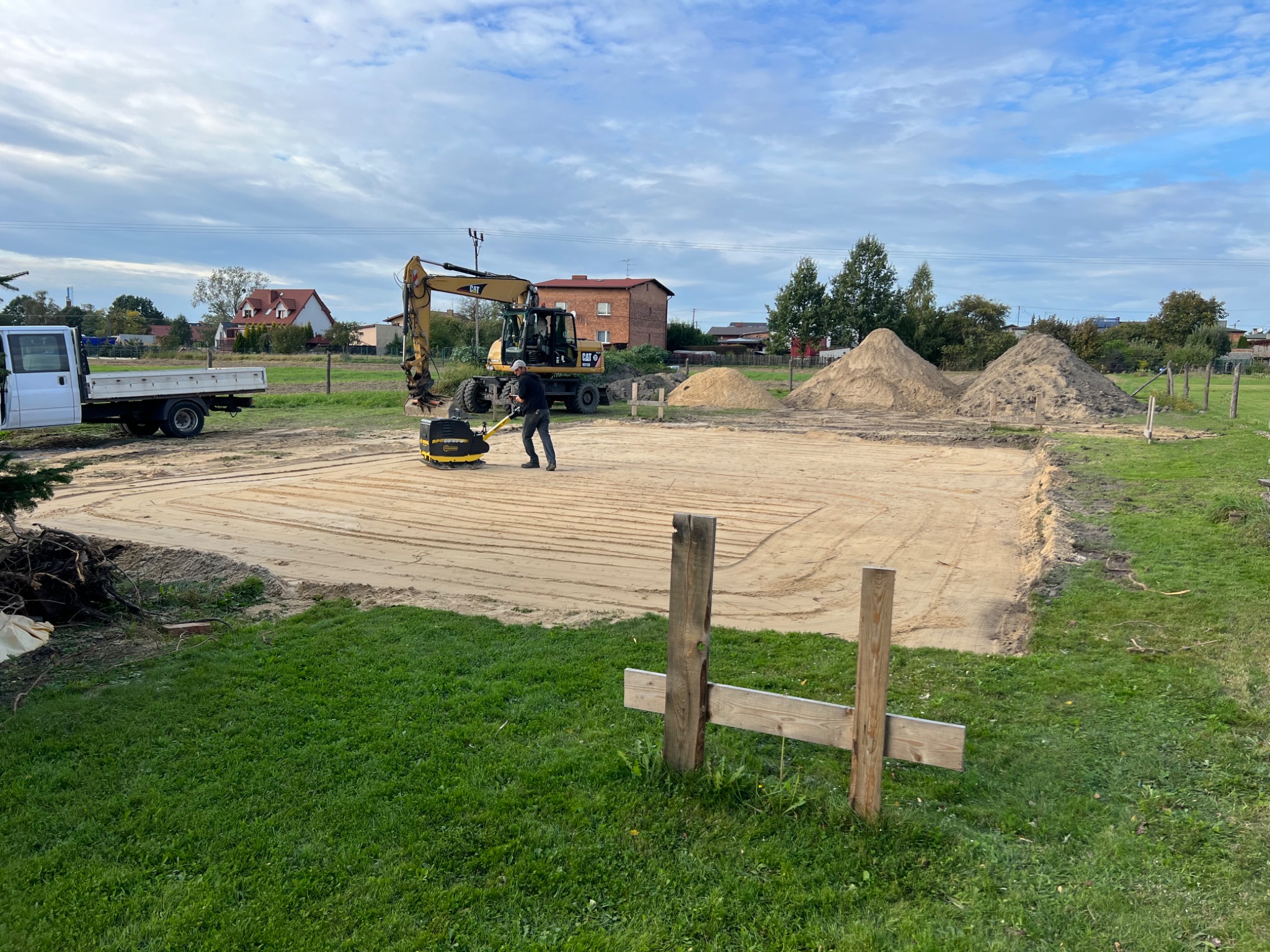 Przygotowanie terenu pod budowę: pracownik zagęszcza piasek zagęszczarką wibracyjną, w tle koparka CAT i hałdy piasku.