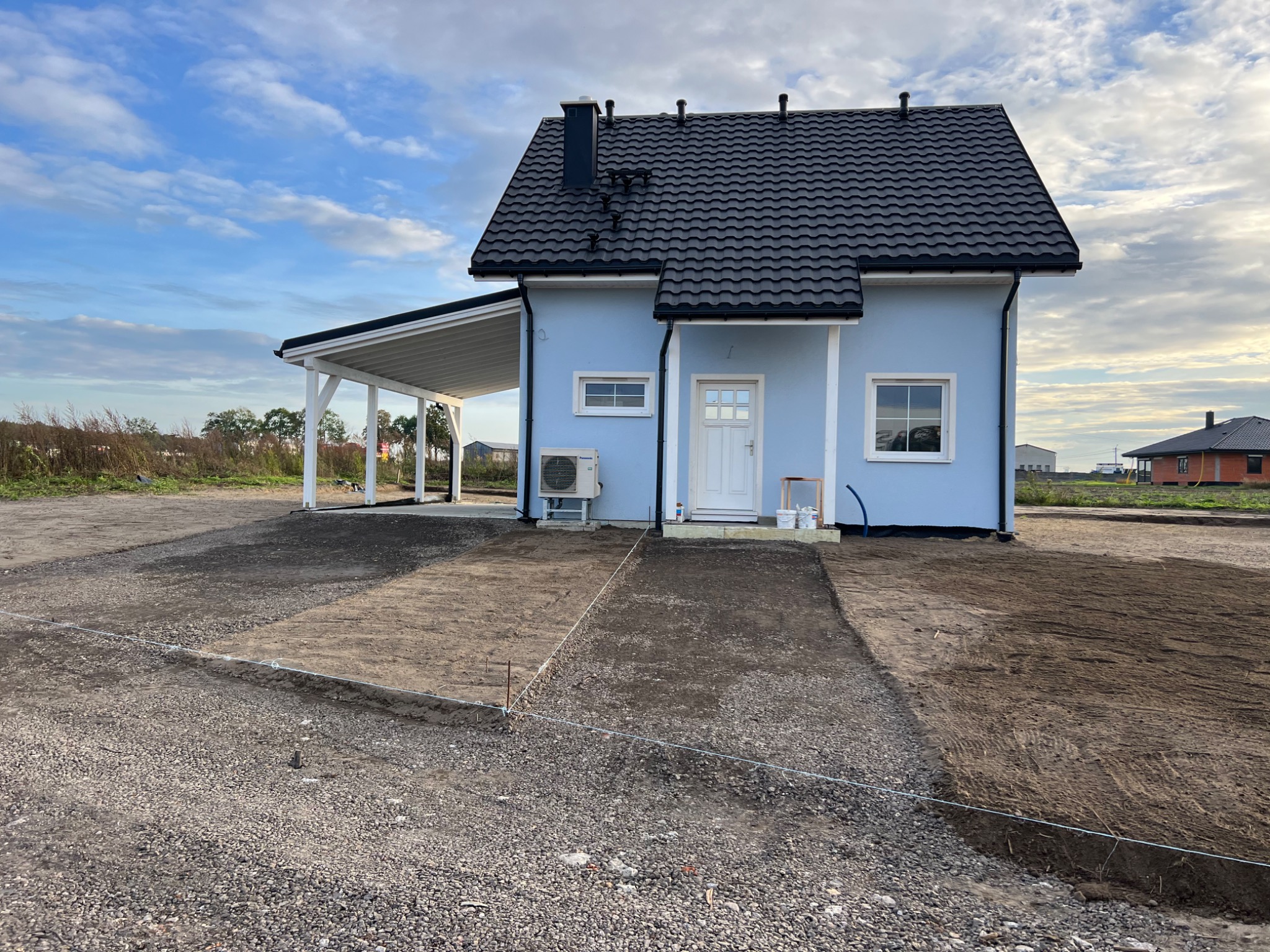 Niebieski dom jednorodzinny z czarnym dachem i białą wiatą garażową w trakcie prac przygotowawczych do zagospodarowania terenu, widoczne wytyczone obrysy przyszłego chodnika z kamienia.