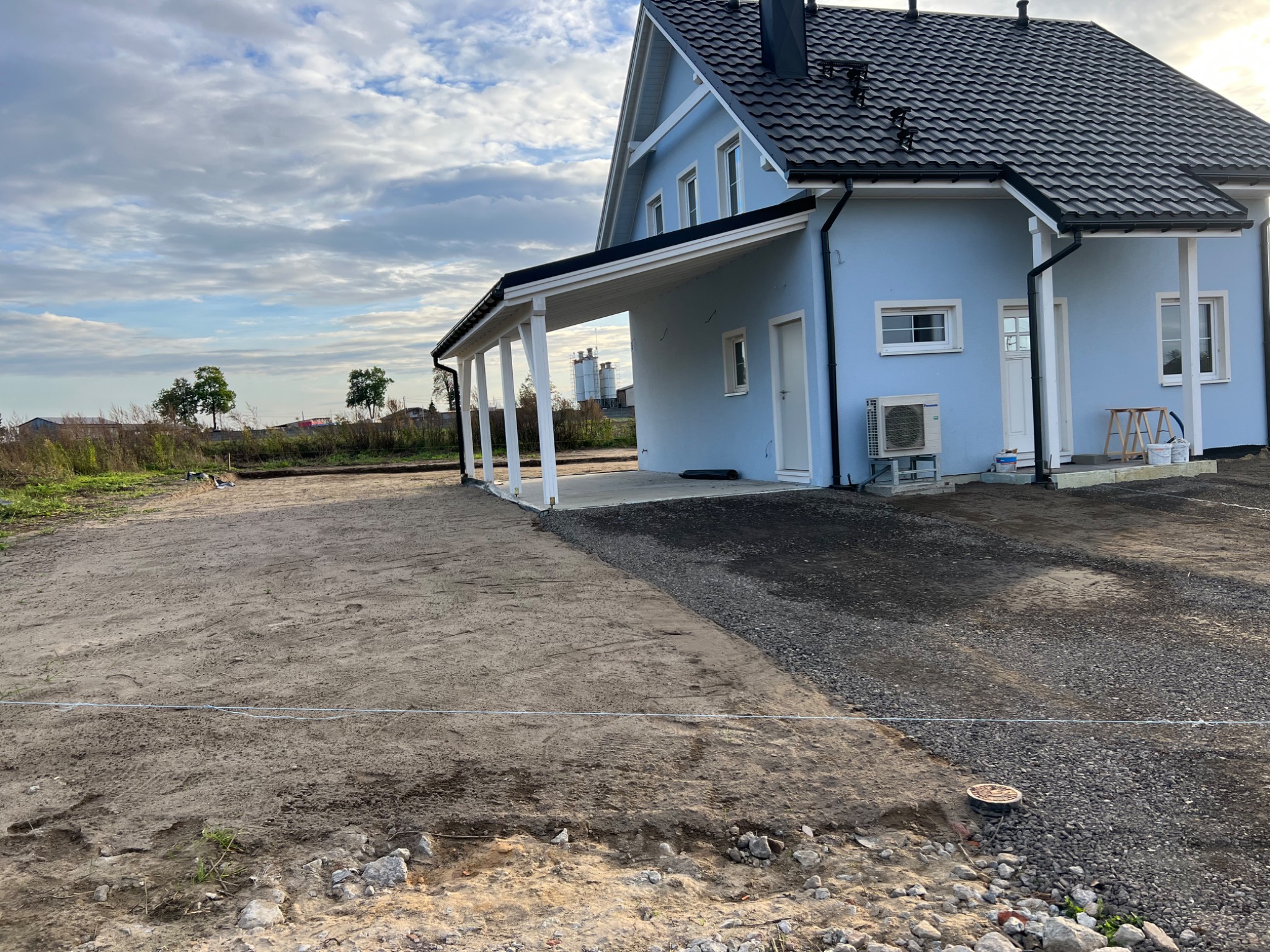 Nierówny teren przygotowany pod budowę, częściowo utwardzony żwirem, z widocznym nowo wybudowanym, jasnoniebieskim domem z czarnym dachem i białymi kolumnami werandy.