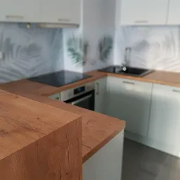 Nowoczesna kuchnia z drewnianym blatem, fronty szafek w odcieniach szarości, backsplash z motywem liści palmowych, płyta indukcyjna i czarny zlew.