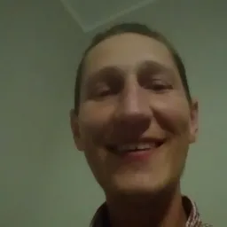 Uśmiechnięty mężczyzna w koszuli w kratę na tle jasnej ściany i fragmentu sufitu. Widoczne zęby i zmarszczki mimiczne.