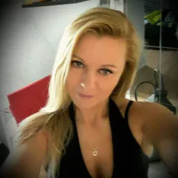 Portret blondwłosej kobiety w czarnej bluzce z wisiorkiem w kształcie serca, z delikatnym uśmiechem i jasnymi oczami, na rozmytym tle wnętrza.