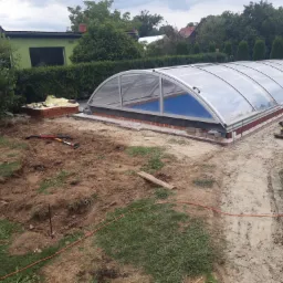 Przygotowanie podłoża pod basen z zadaszeniem, widoczne narzędzia i ubijarka, częściowo ułożona kostka wokół basenu.