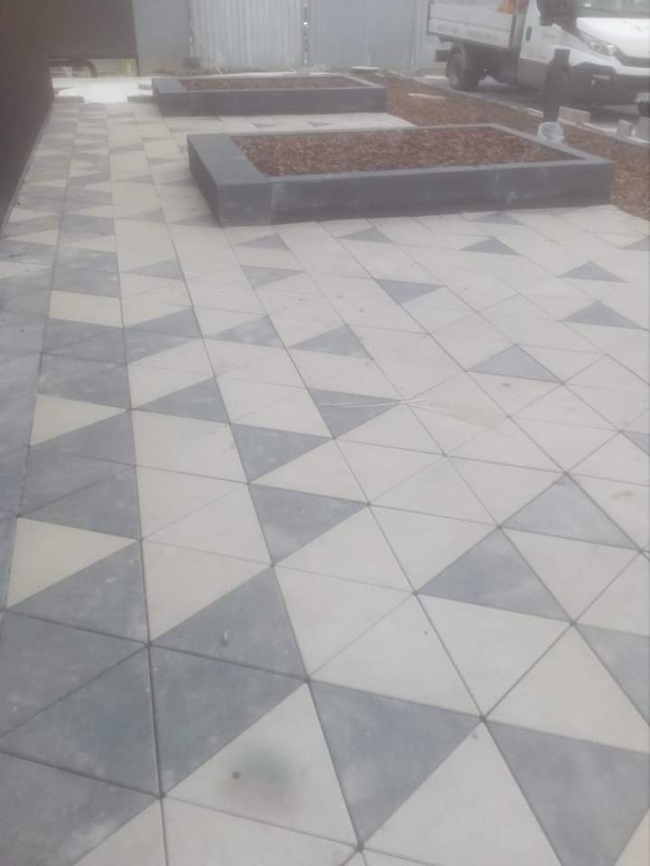Nawierzchnia z trójkątnych kostek brukowych w odcieniach szarości i bieli, z donicami wypełnionymi korą w tle i samochodem dostawczym w oddali.