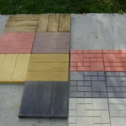 Różnorodne wzory kostki betonowej ułożone na betonowej powierzchni, prezentujące imitacje drewna, kamienia łupanego i cegły w odcieniach brązu, różu, szarości i beżu.