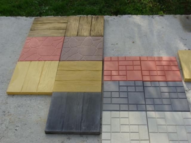 Różnorodne wzory kostki betonowej ułożone na betonowej powierzchni, prezentujące imitacje drewna, kamienia łupanego i cegły w odcieniach brązu, różu, szarości i beżu.