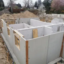 Wznoszenie fundamentów budynku z betonu i bloczków, widoczne szalunki okienne, ziemia wokół fundamentów, drzewa w tle.